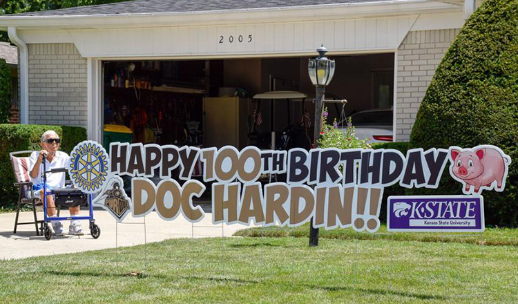 Doc Hardin sits in his driveway next to a large sign that says Happy 100th Birthday Doc Hardin with a Rotary Club symbol, Purdue Boilermaker Special logo, and Kansas State sign. There is also a pig to represent his farm and veterinary career.