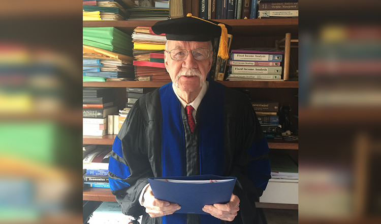 James Franklin Sharp stands in his PhD cap and gown in front of a bookshelf in his NYC apartment
