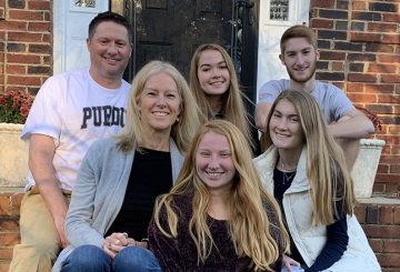 Volpp Family. Two parents, three teenage daughters and one college age son sit together on the front steps of a brick building, smiling, and looking directly at the camera.