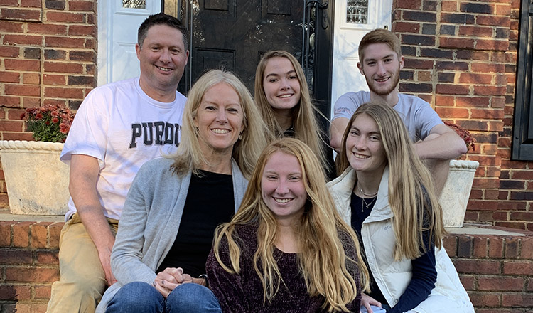Volpp Family. Two parents, three teenage daughters and one college age son sit together on the front steps of a brick building, smiling, and looking directly at the camera.